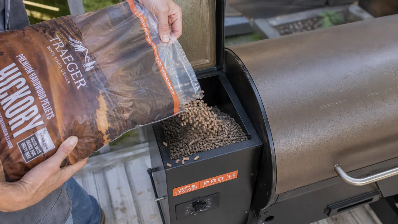 Close-up of Traeger Pro 34 pellet hopper lid open showing wood pellets being poured in from a bag, hand visible